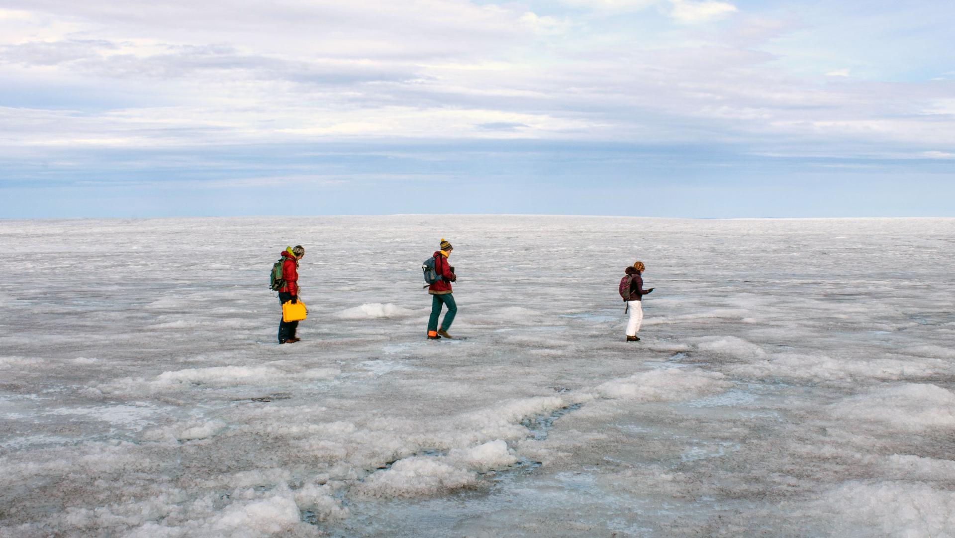 Three people walk across ground covered in ice and snow as the horizon stretches behind them.
