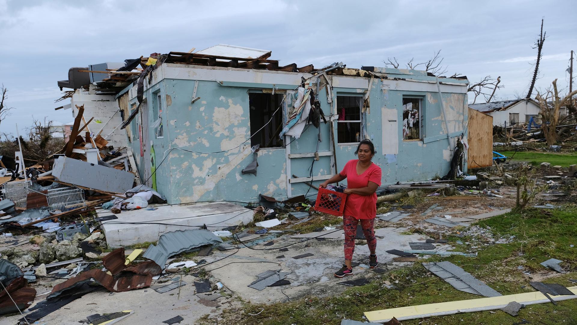 A woman is shown wearing a red shirt and walking with a crate next to a heavily damaged home.