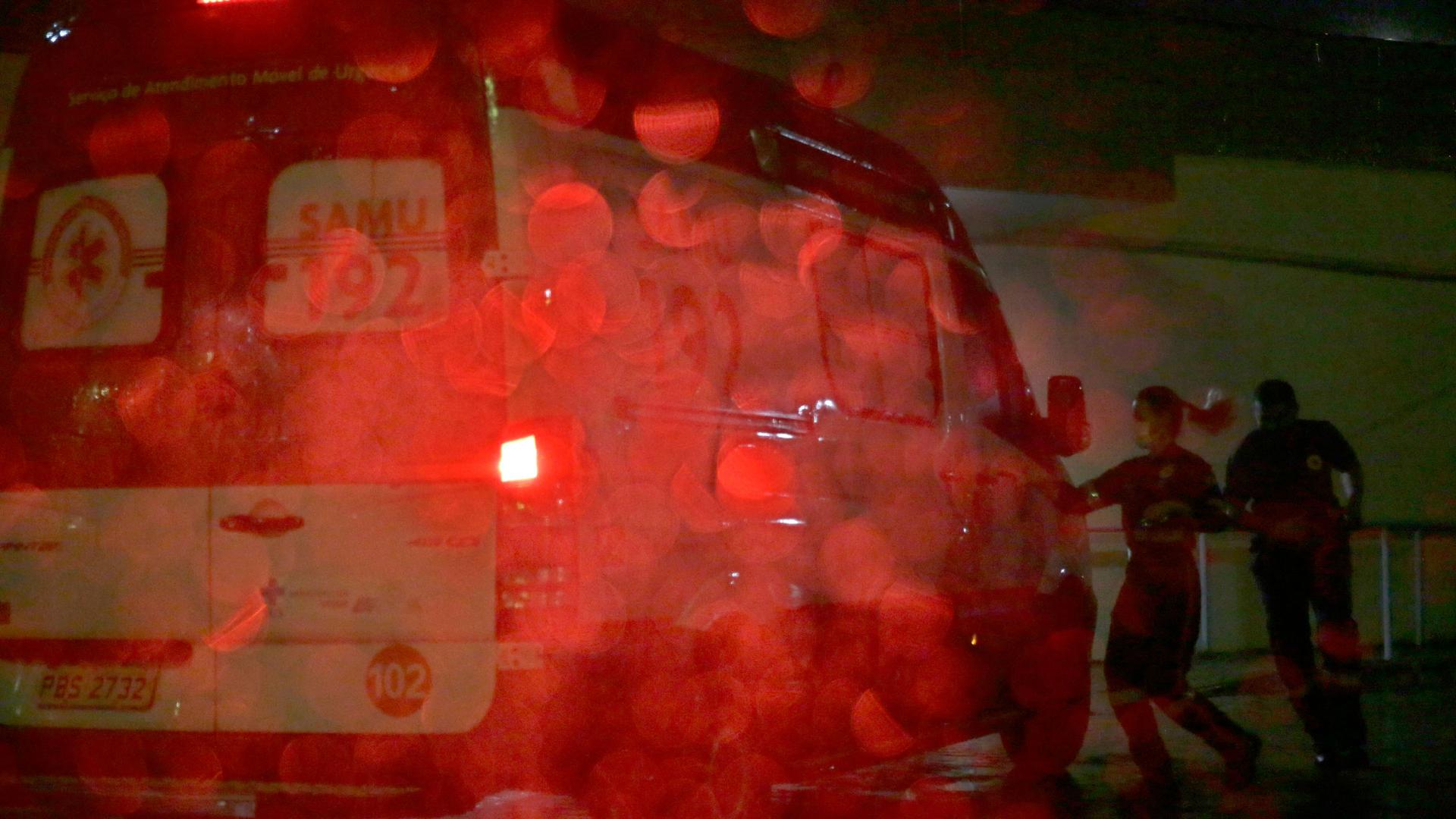 Two people are shown in shadow running toward an ambulance with red brake lights on in the rain.