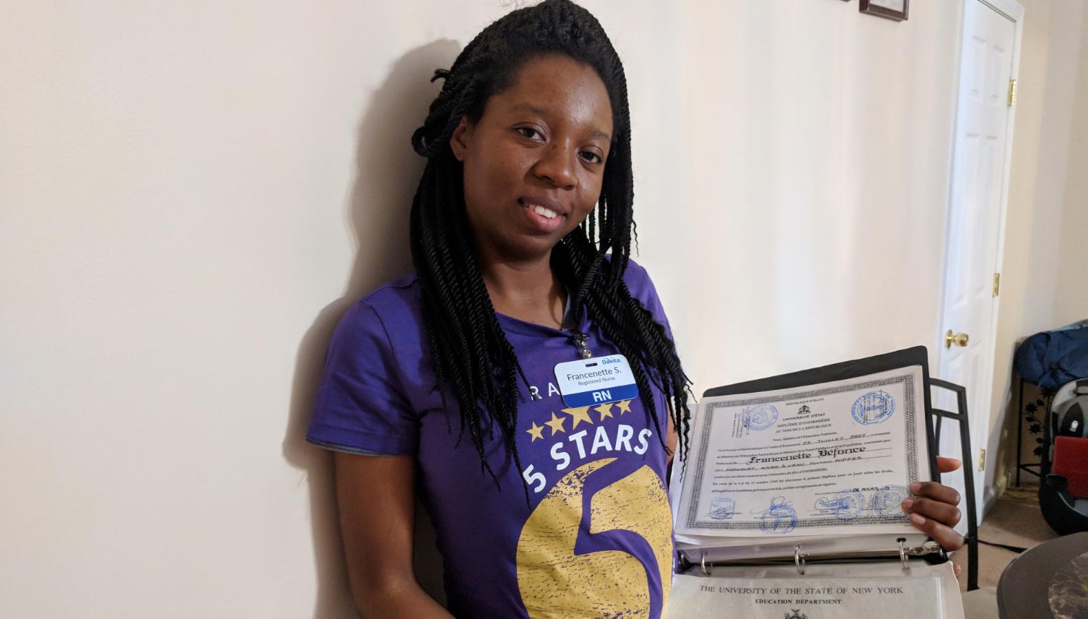 Woman against white wall holding up certificate that shows nursing credentials