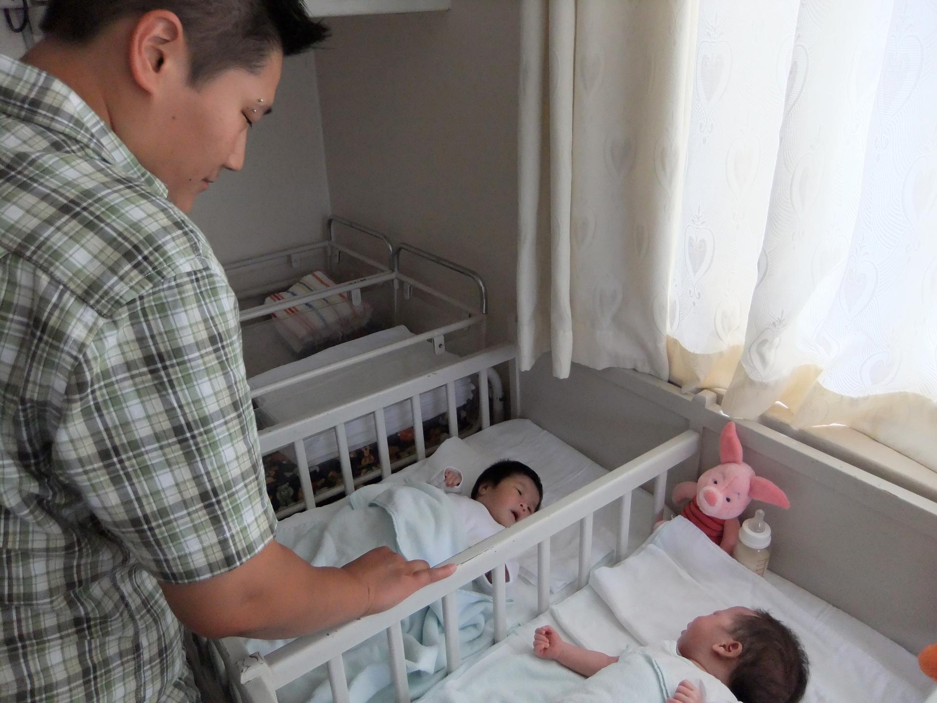 A woman stands over two cribs with babies in them.