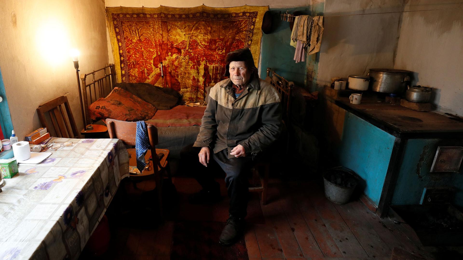 Vitaliy Kudla is shown wearing a two-tone jacket and hat while sitting in a small room with a bed behind him.