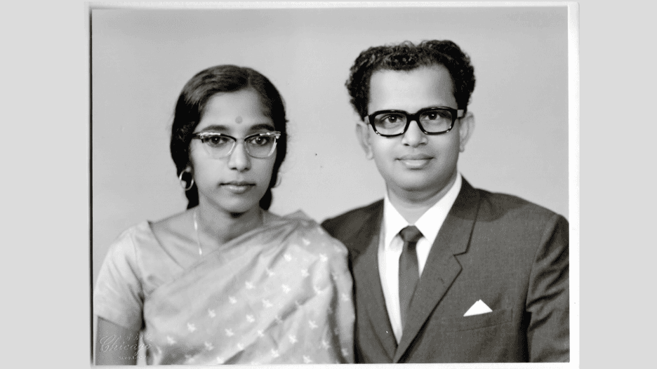 An Indian woman in a sari and glasses and an Indian man in a suit and glasses sit for a portrait.