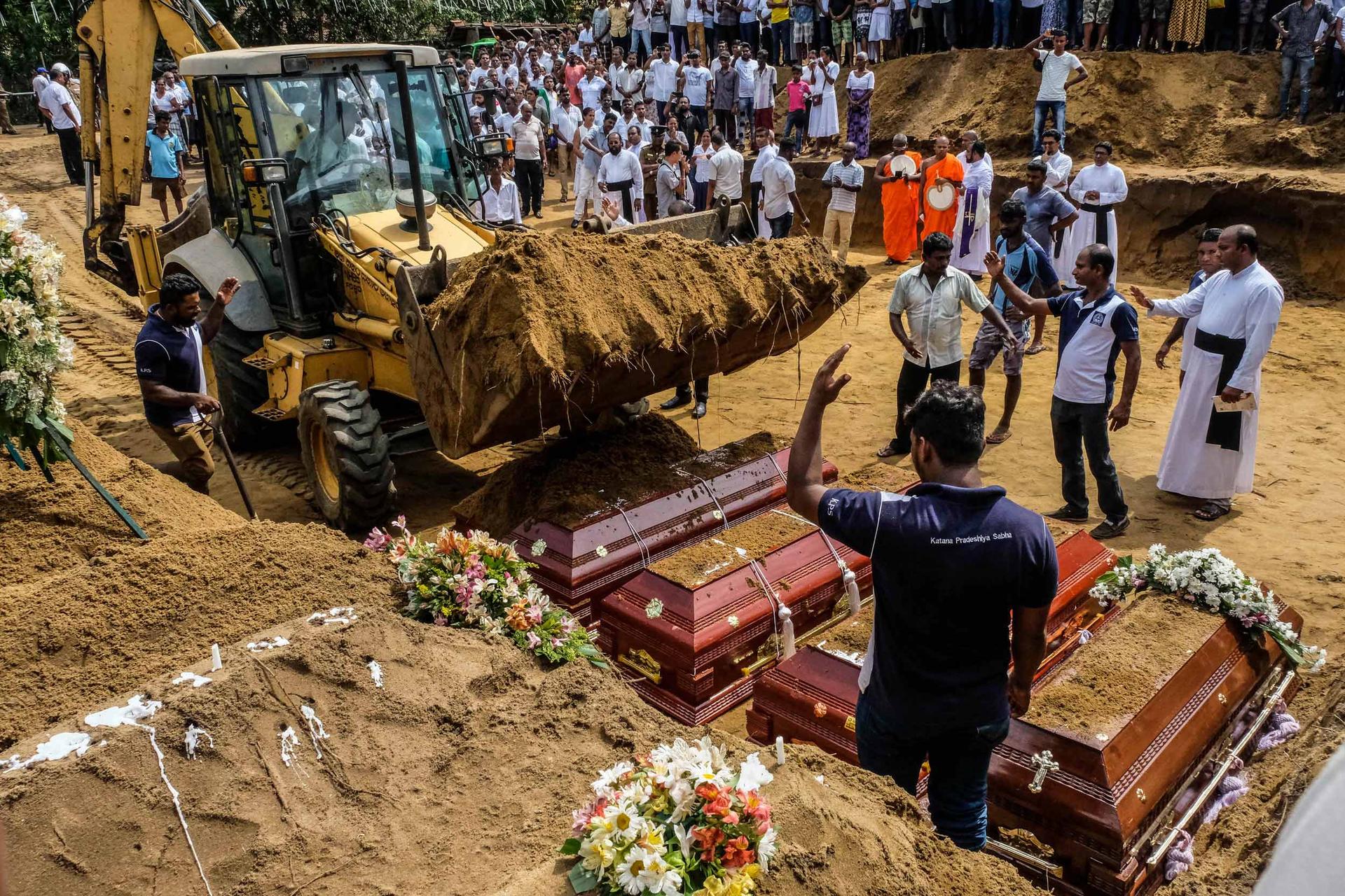 A man waves a backhoe as it lifts dirt to drop onto four caskets laid out in a row