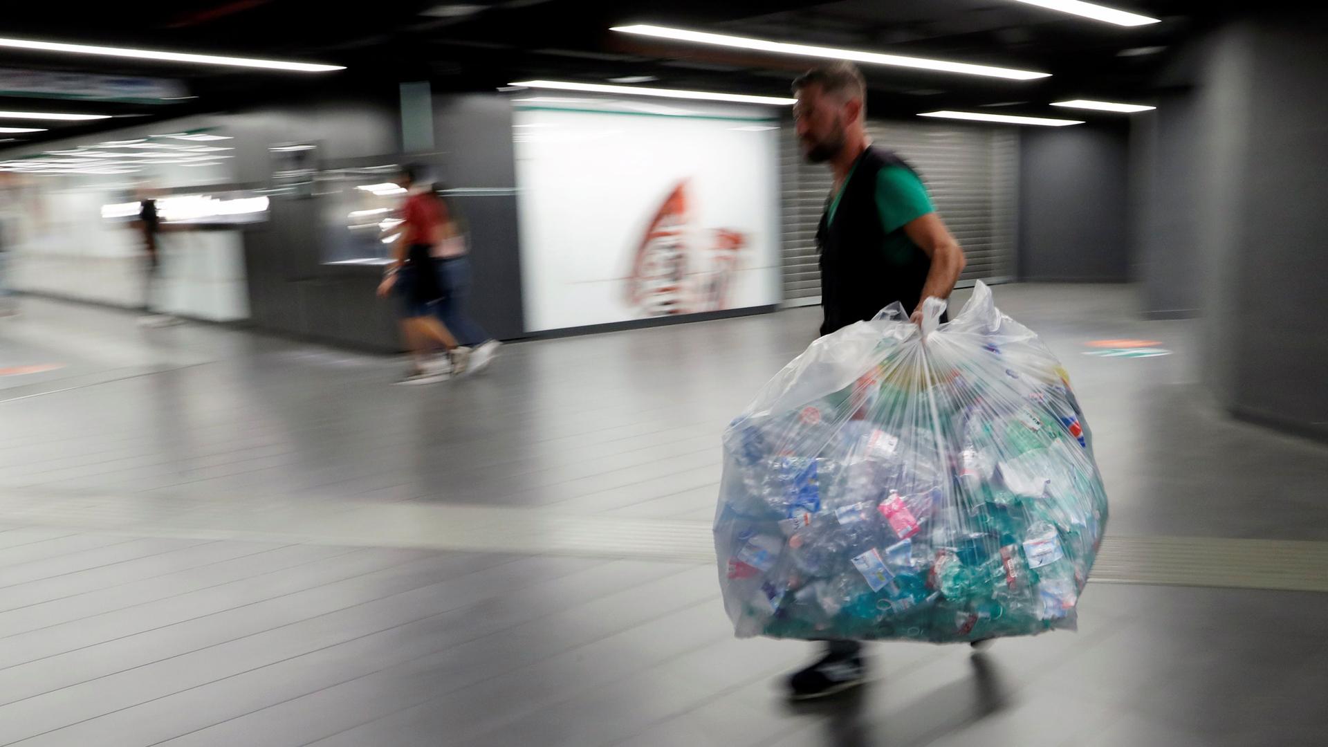 A man is shown wearing a dark-color vest and carrying a large garbage bag full of plastic bottles.