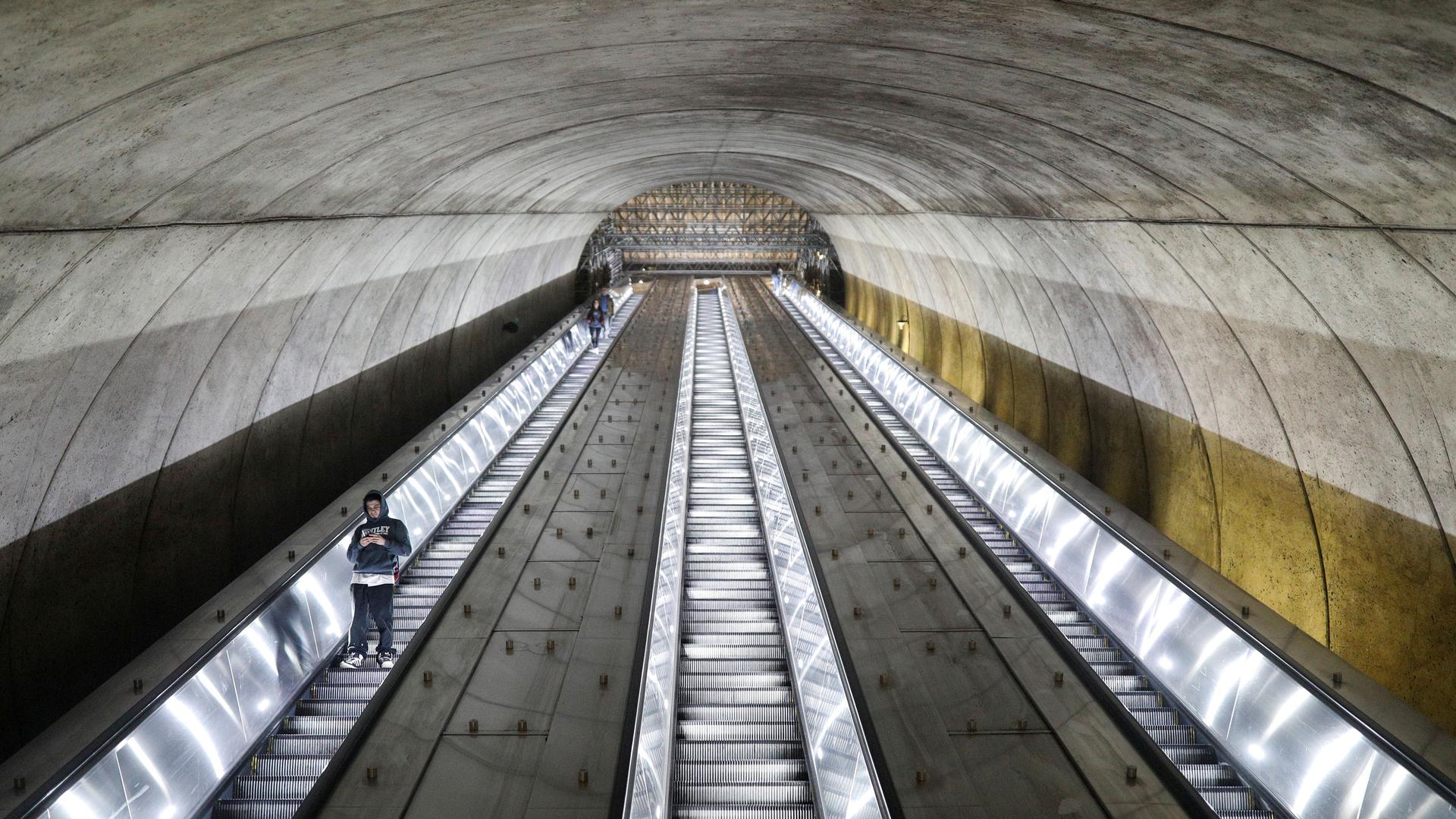 A long escalator is shown nearly empty with a person riding down and looking at their phone.