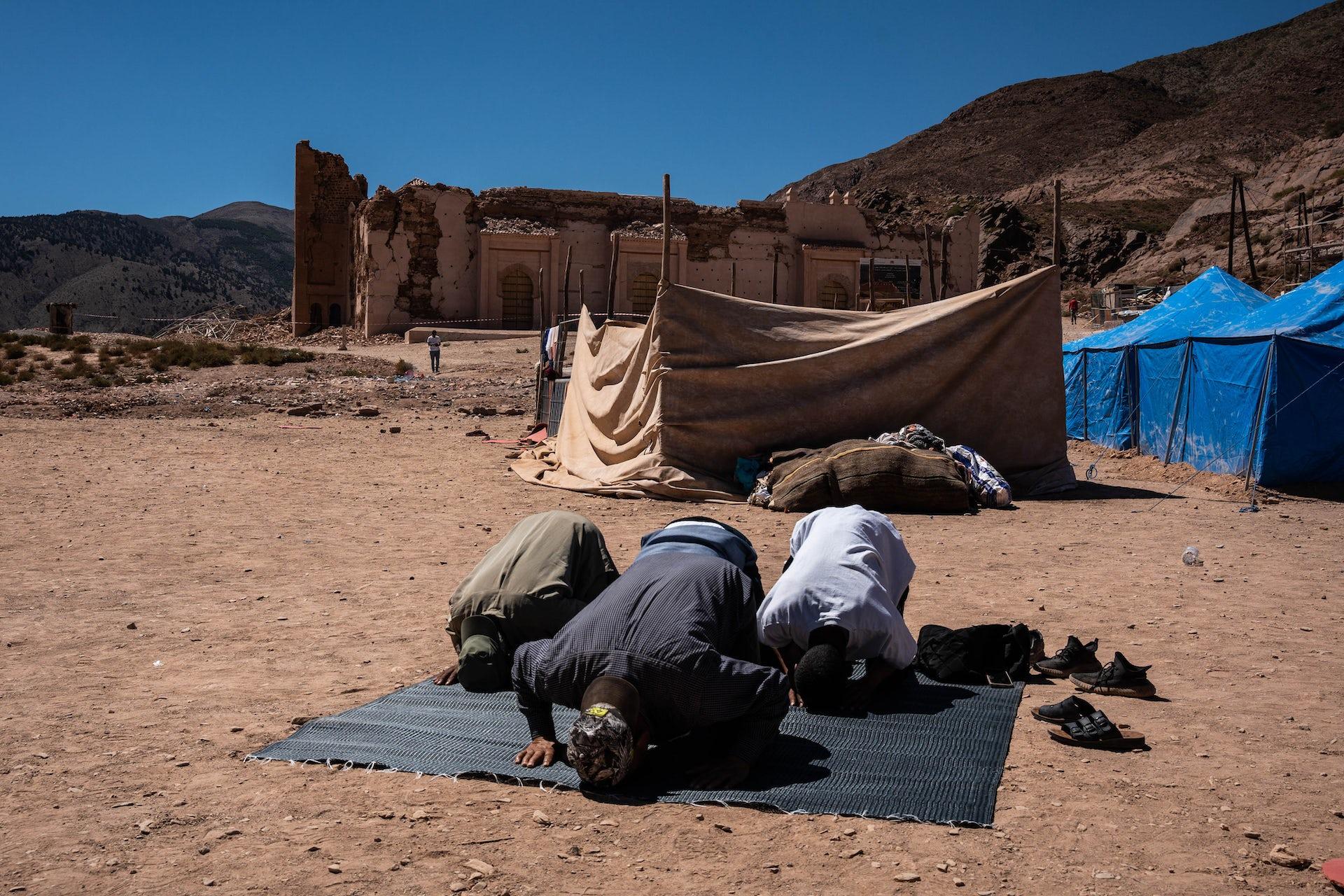 Three people prostrating in prayer on a mat. In the backdrop are mountains and a damaged ancient brick structure.