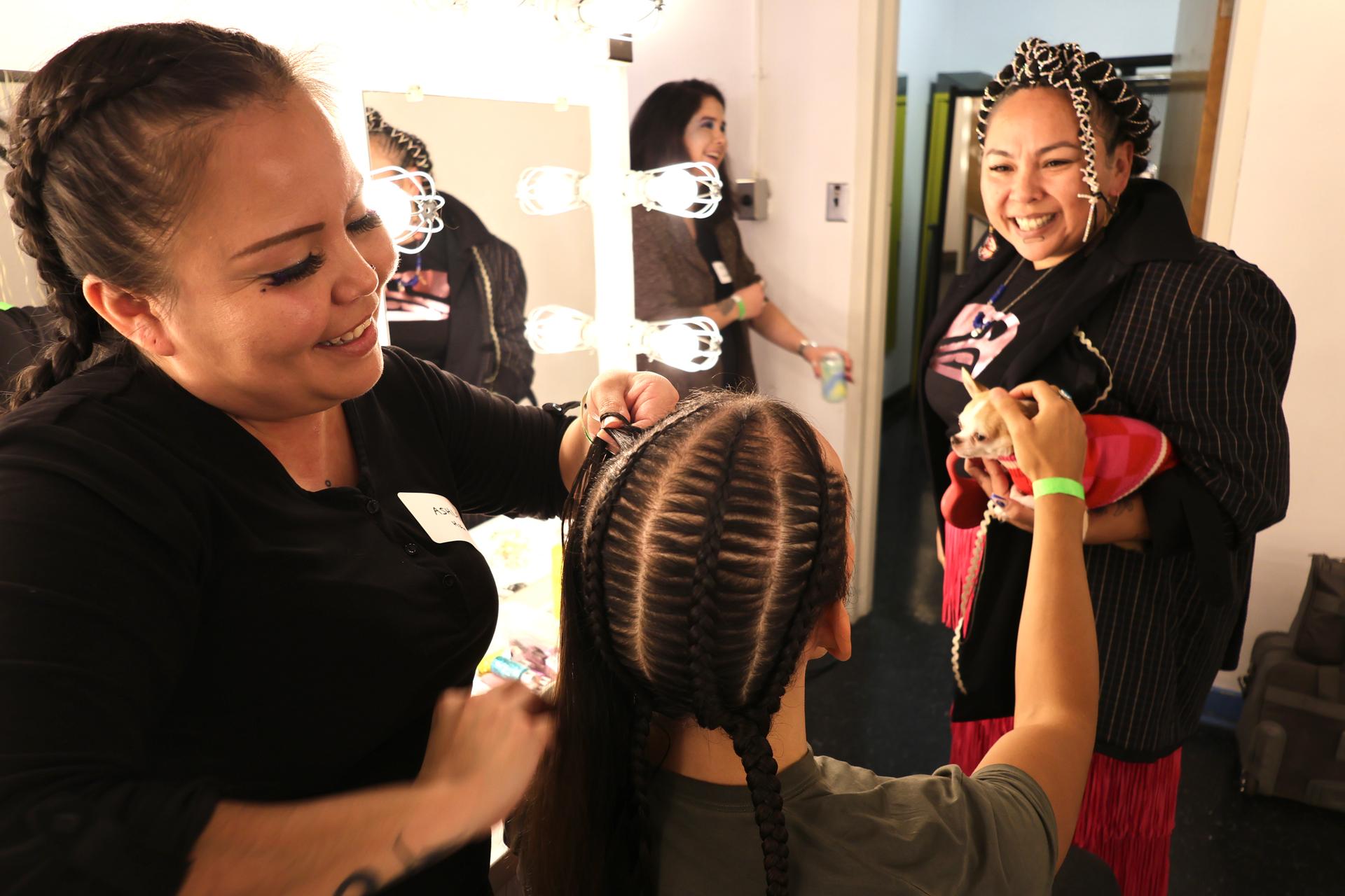 Hair stylist Ashley Jack (left) braids hair on model Ricky-Lee Watts (center) before Indigenous Futures streetwear fashion night. Lauraleigh Paul (right) smiles as she holds her dog.