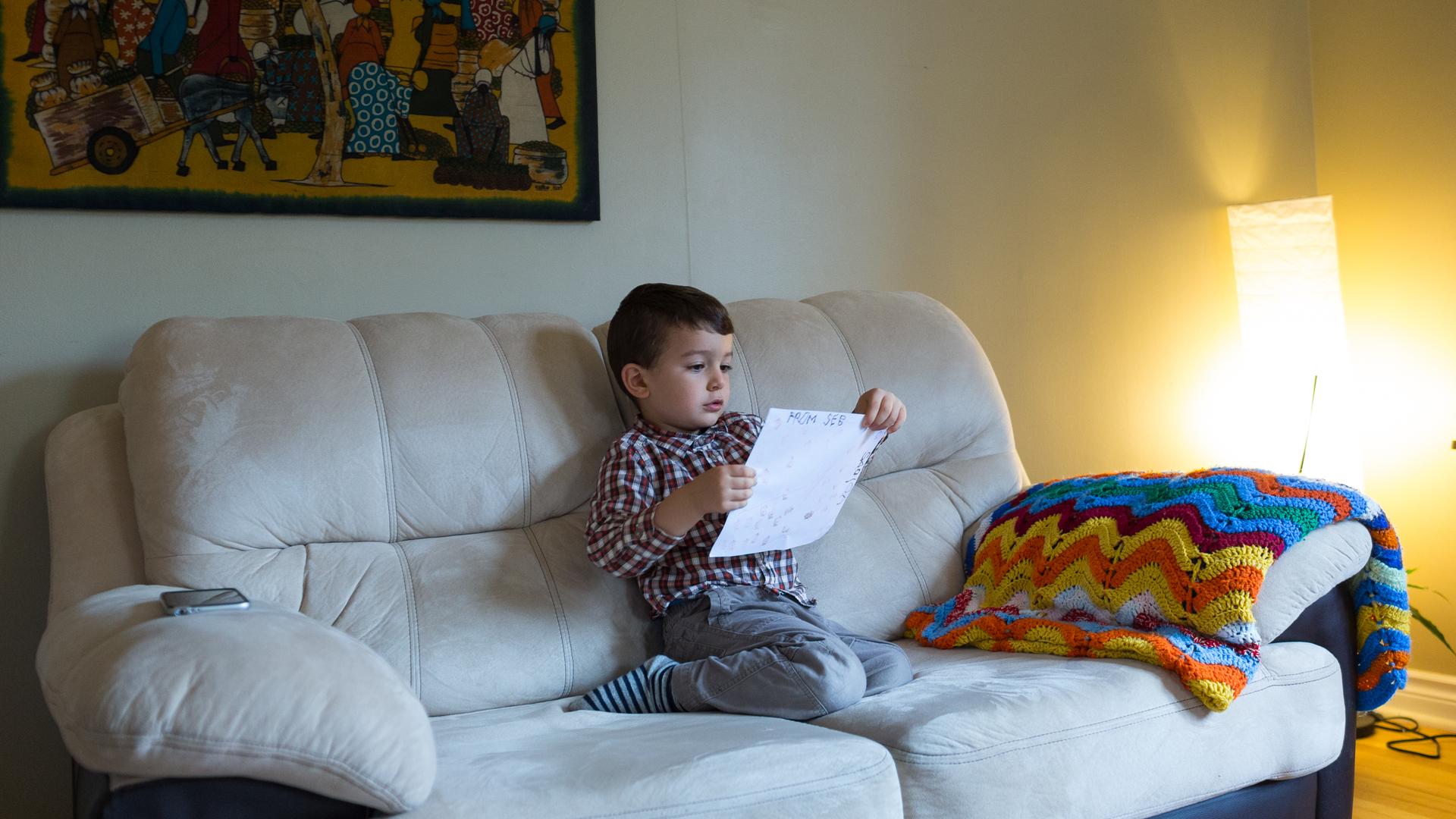 Boy sitting on cream sofa holding child's drawing, looking at it