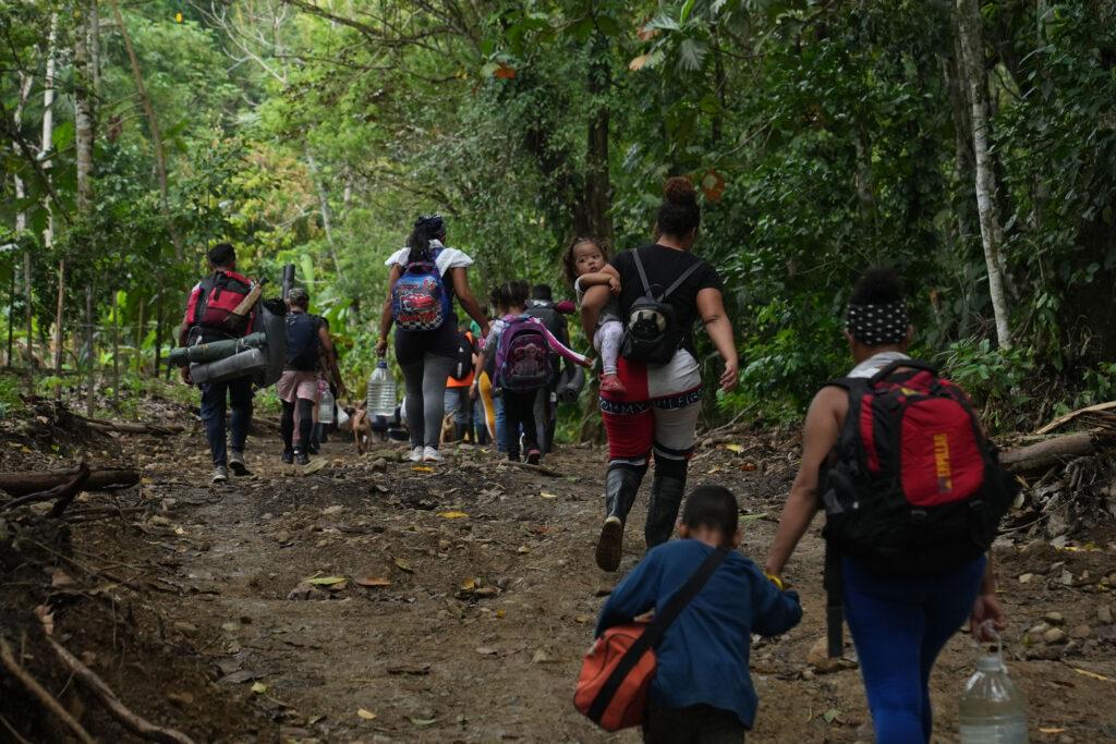 People walk with children through a forested area carrying backpacks and water bottles.