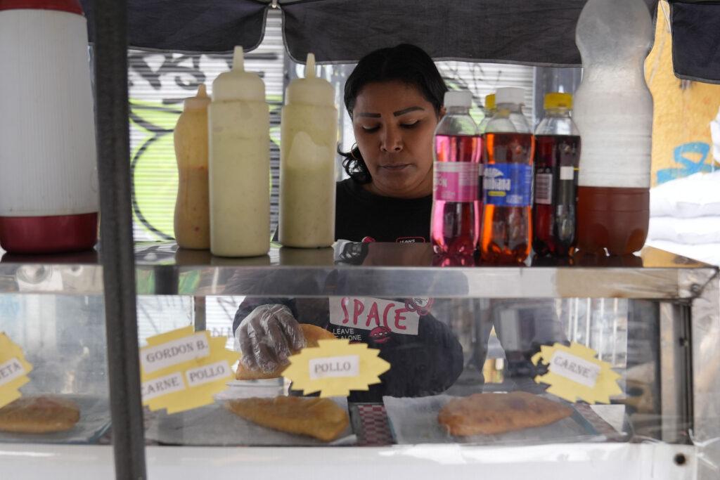 A woman stands behind a glass counter holding an empanada in her gloved hand.