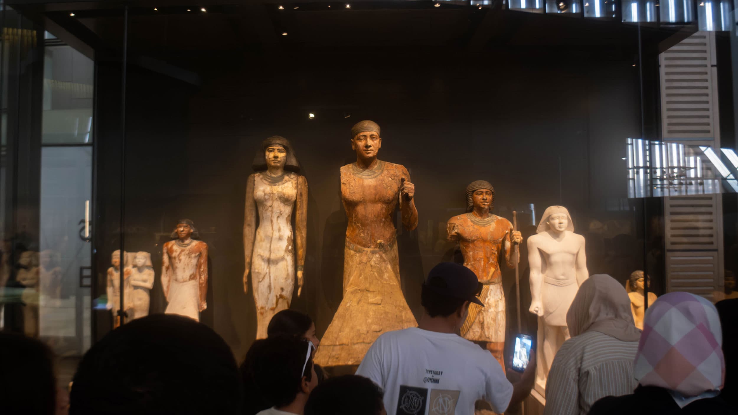 A group of people observing ancient Egyptian statues displayed in a museum exhibit, with the statues positioned behind a glass case.