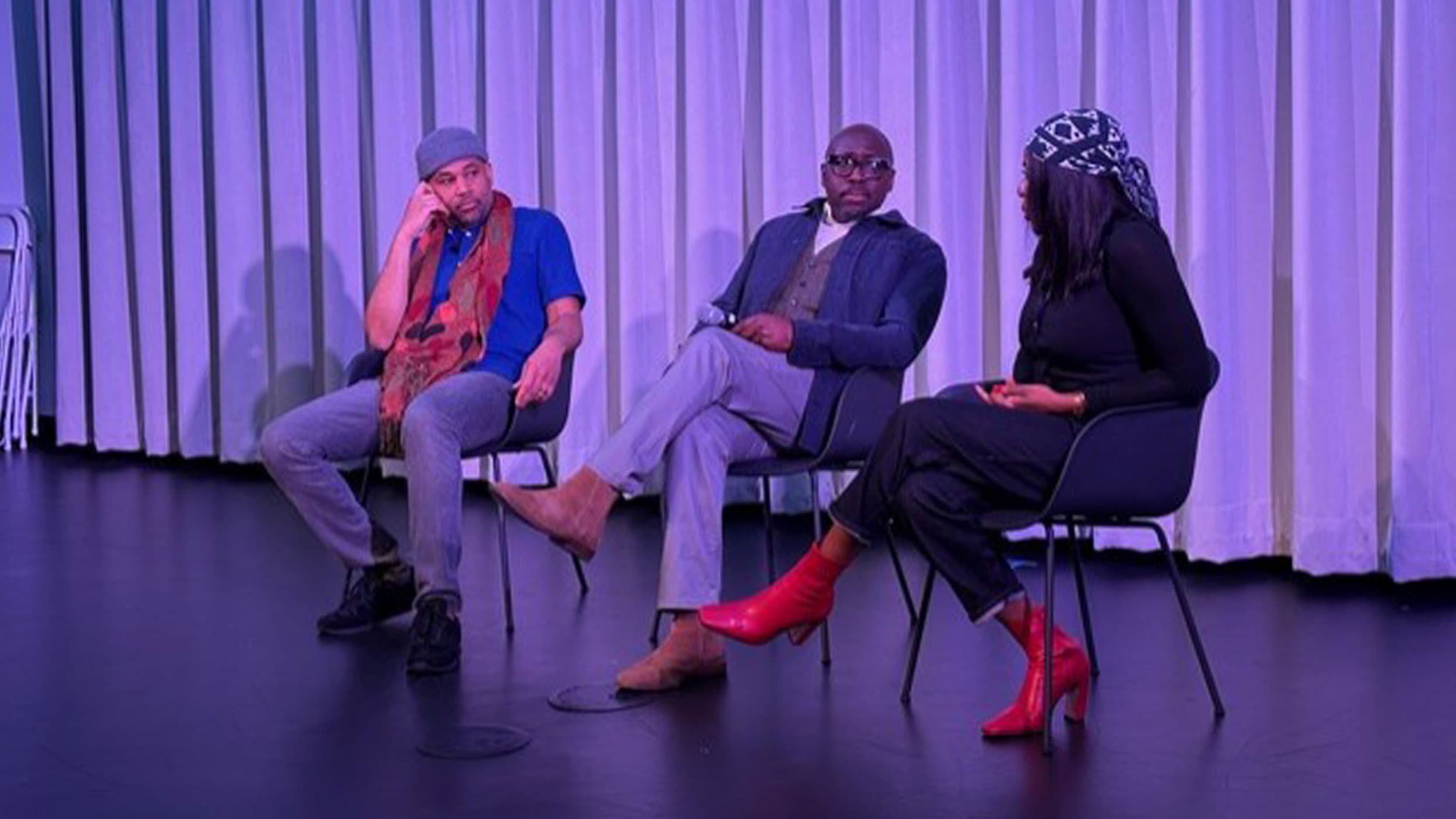 Three people seated in chairs on a stage in front of a curtain, engaged in a discussion, with one man holding a microphone and the woman wearing red boots.