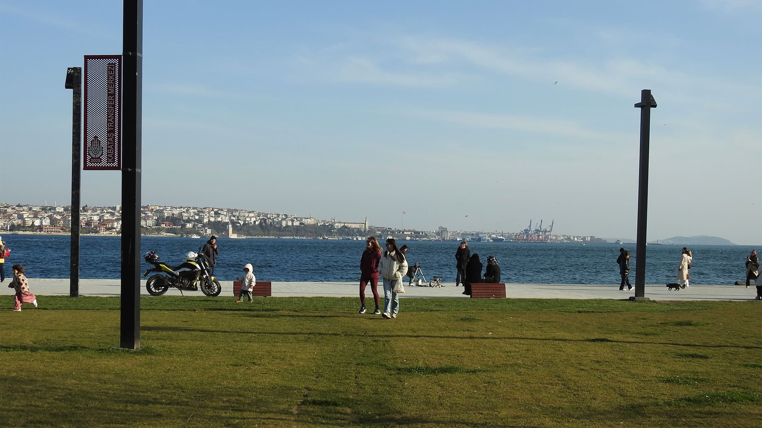 People strolling along a seaside promenade with a cityscape and industrial port visible in the background. A motorcycle is parked near a walkway, and children are playing on the grass in the foreground.