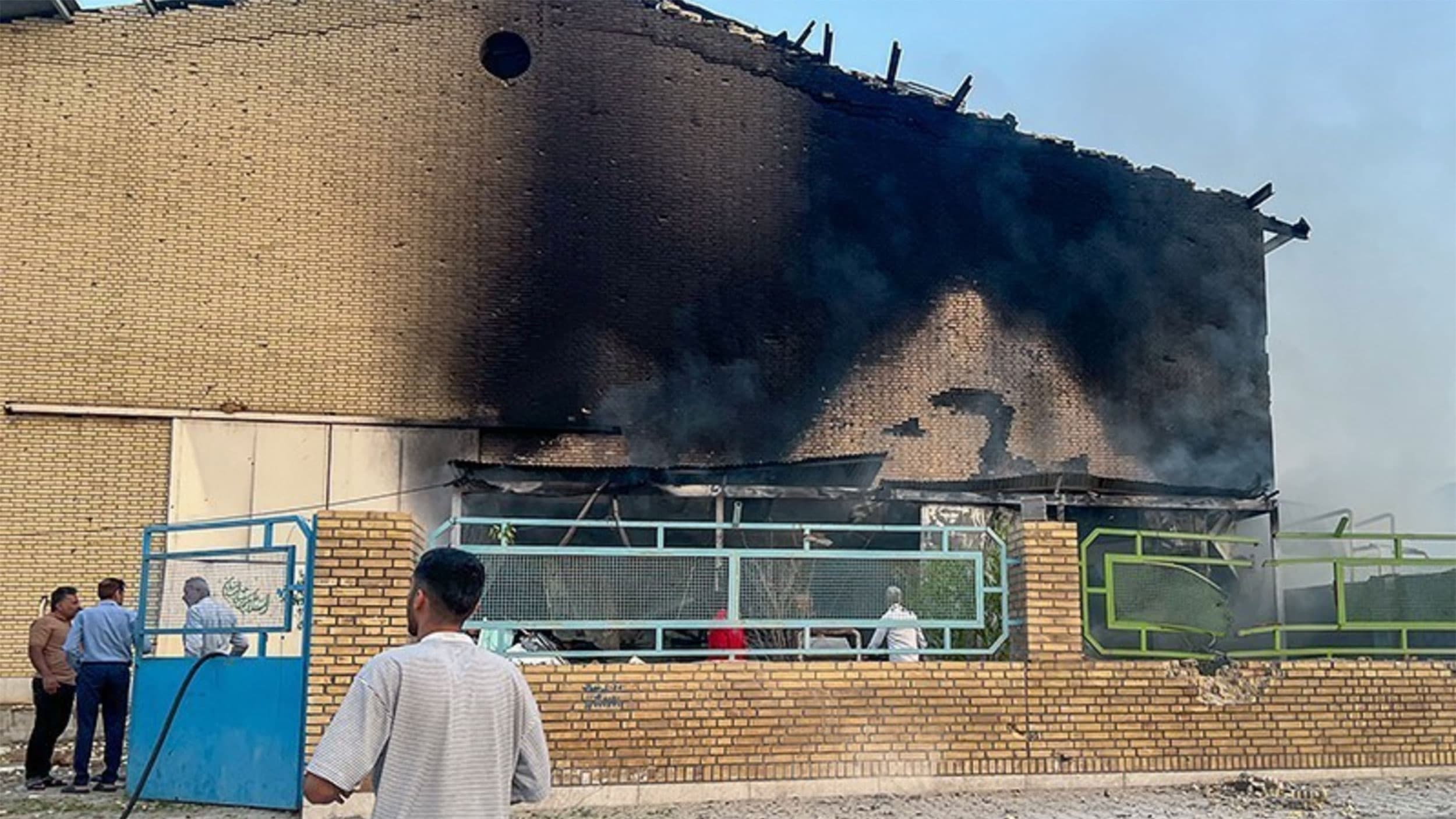 A damaged brick building with blackened marks from a fire, surrounded by people inspecting the site; a blue gate is open in front.
