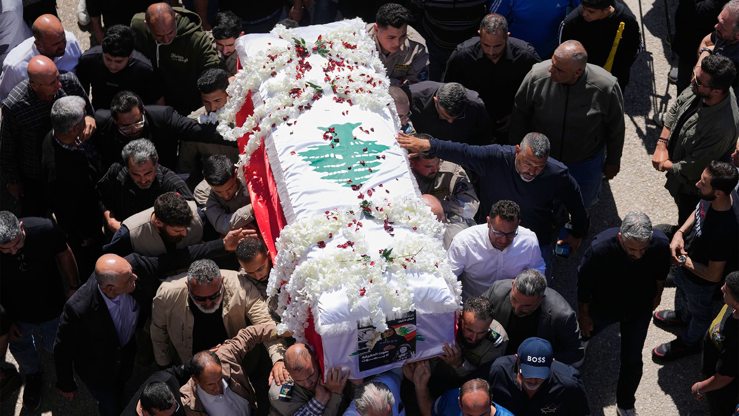 A group of people carrying a coffin draped in a flag with floral decorations, surrounded by a crowd of onlookers.