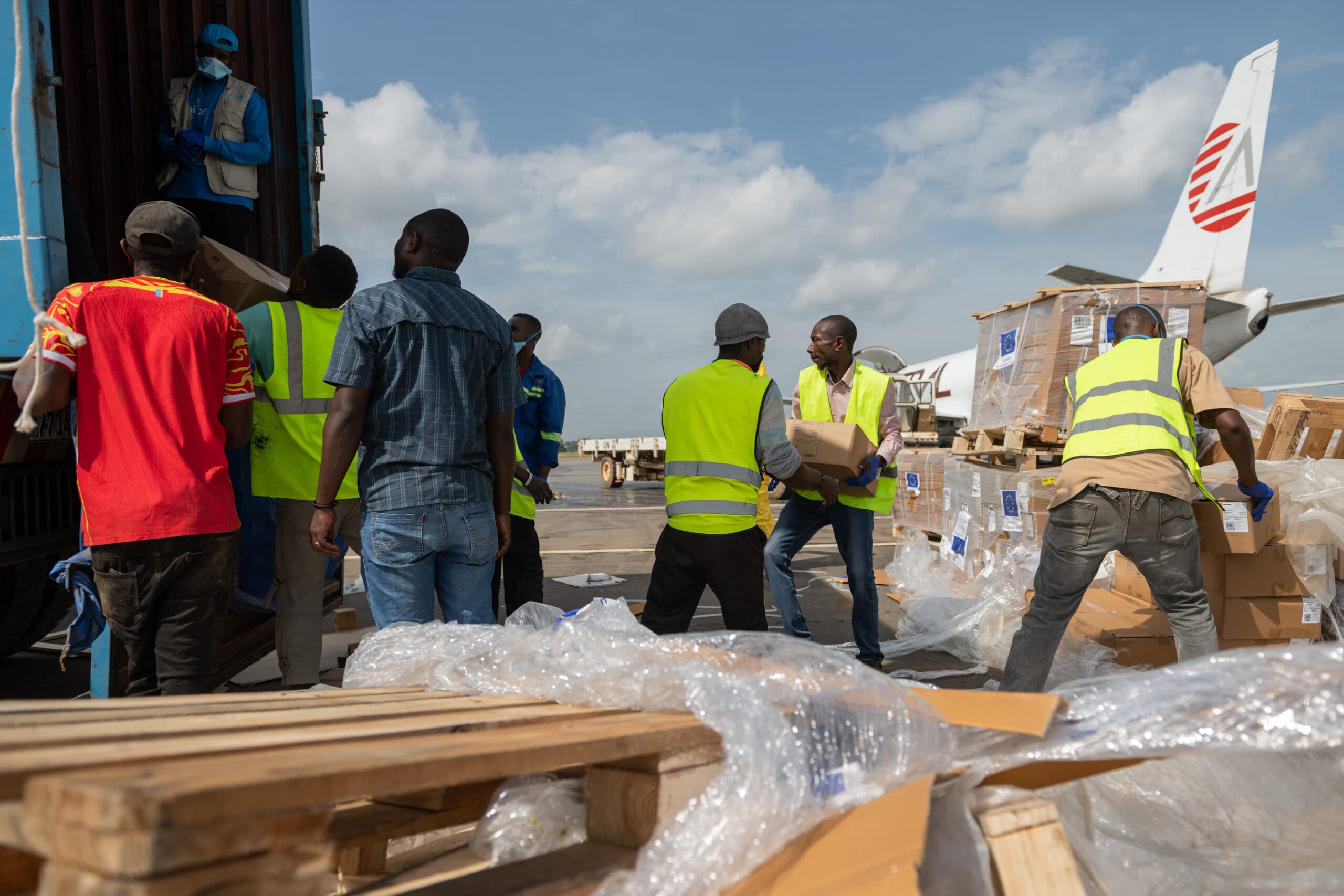 Workers in bright vests unload cargo from a plane onto a tarmac, surrounded by boxes and pallets.