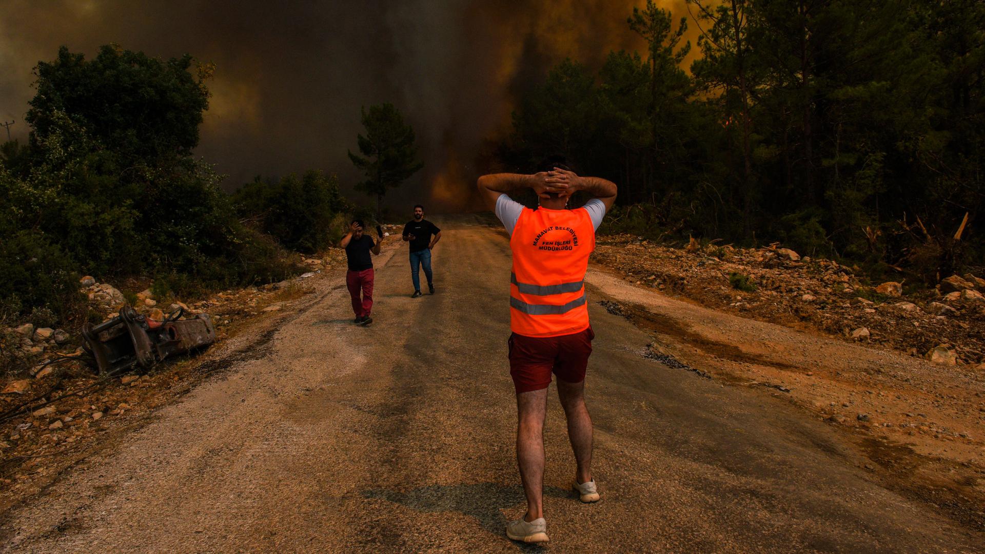 A man is shown with his back to the camera and facing a large wildfire with two other people running away from the fire.