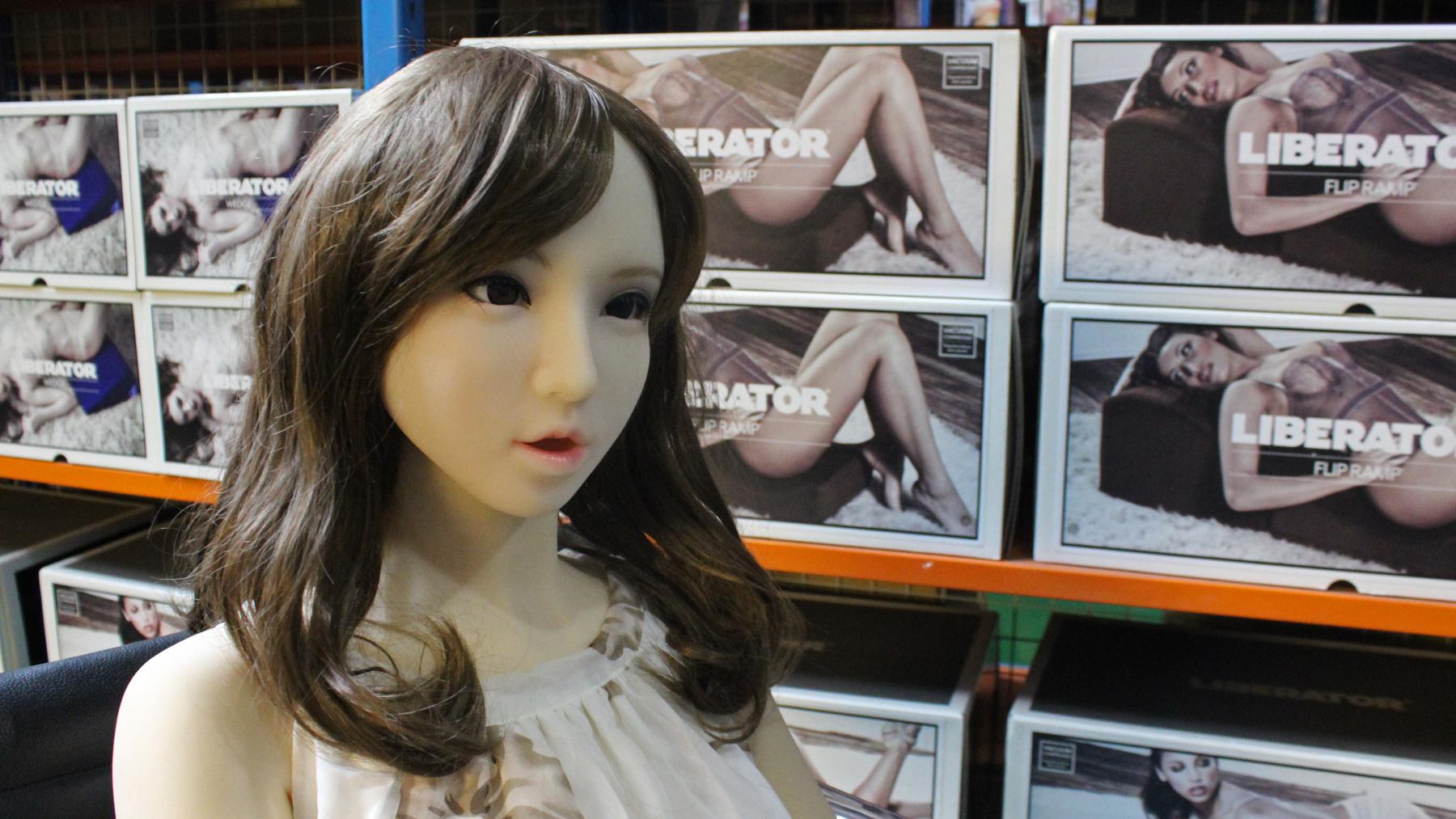 A plastic sex doll with brown wig sits in an office chair