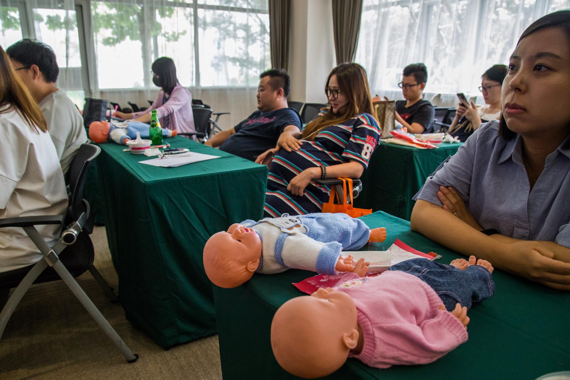 A visibly pregnant woman sits in a parenting class with dolls on the table.