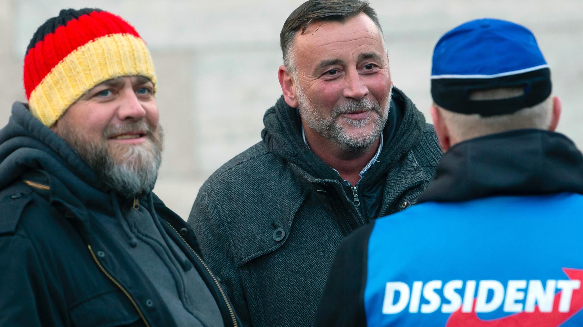 Three white men stand together outside. One wears a hat with German flag colors. One's blue shirt back reads "Dissident."