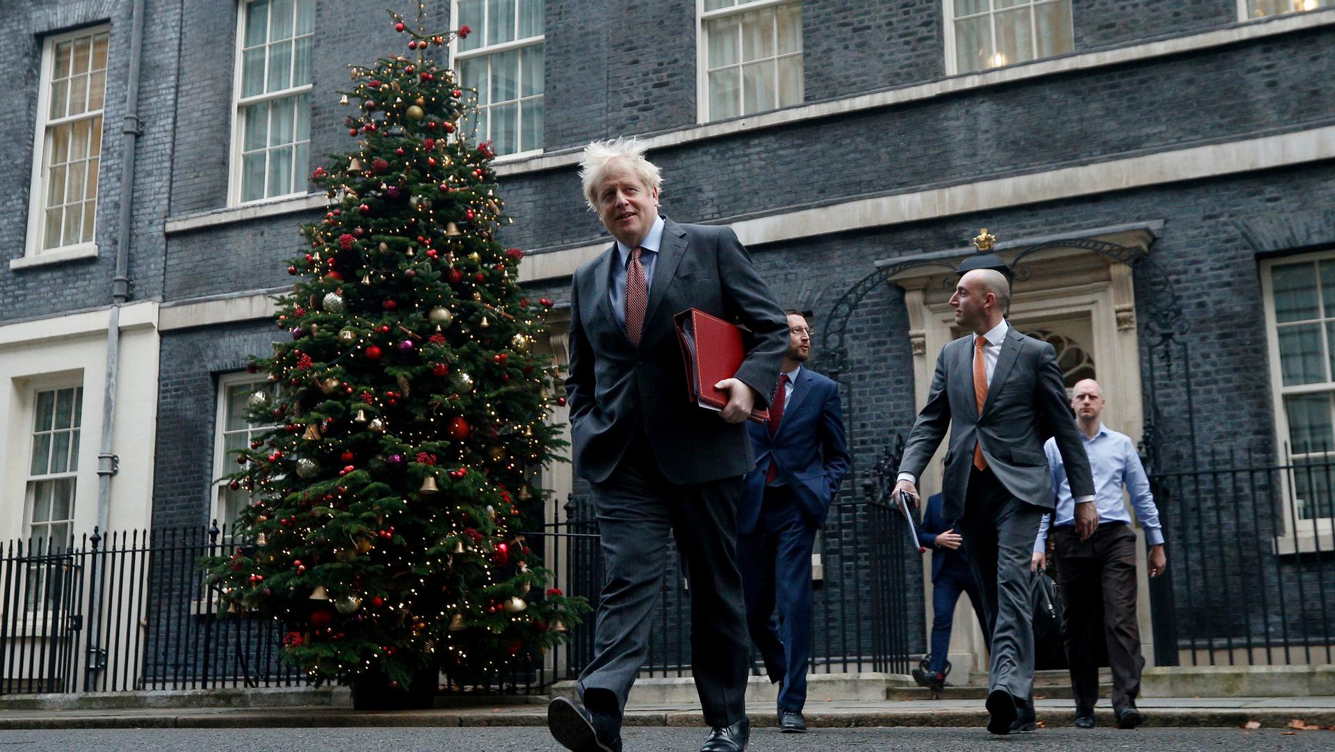 British Prime Minister Boris Johnson is shown wearing a suit and walking with a red folder under his arm and a large Christmas tree in the background.