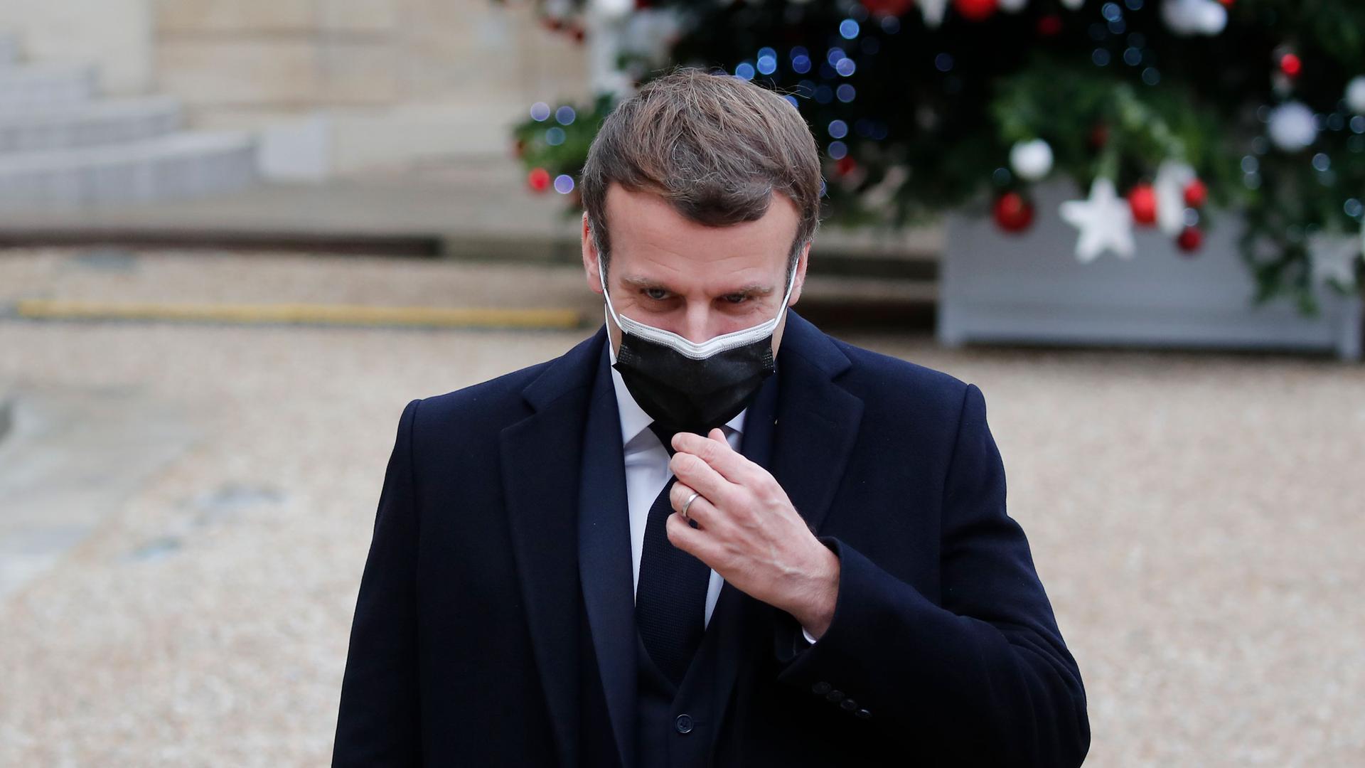 French President Emmanuel Macron is shown wearing a face mask and dark suit while standing outside.