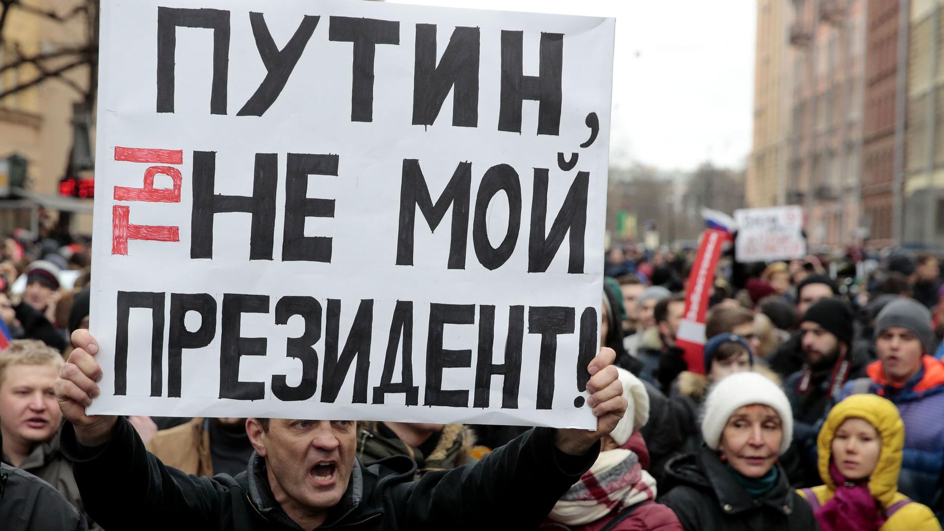 A man holds a white sign with lettering in Russian. He is surrounded by dozens of others. The sign says "Putin, you are not my president."