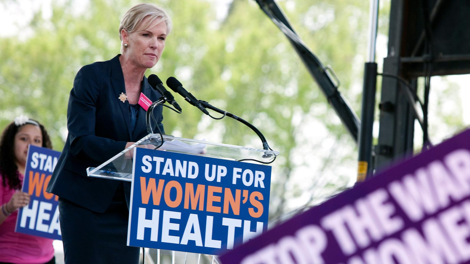A blond woman is speaking into a microphone at a podium with a sign on the front that says "Stand up for women"