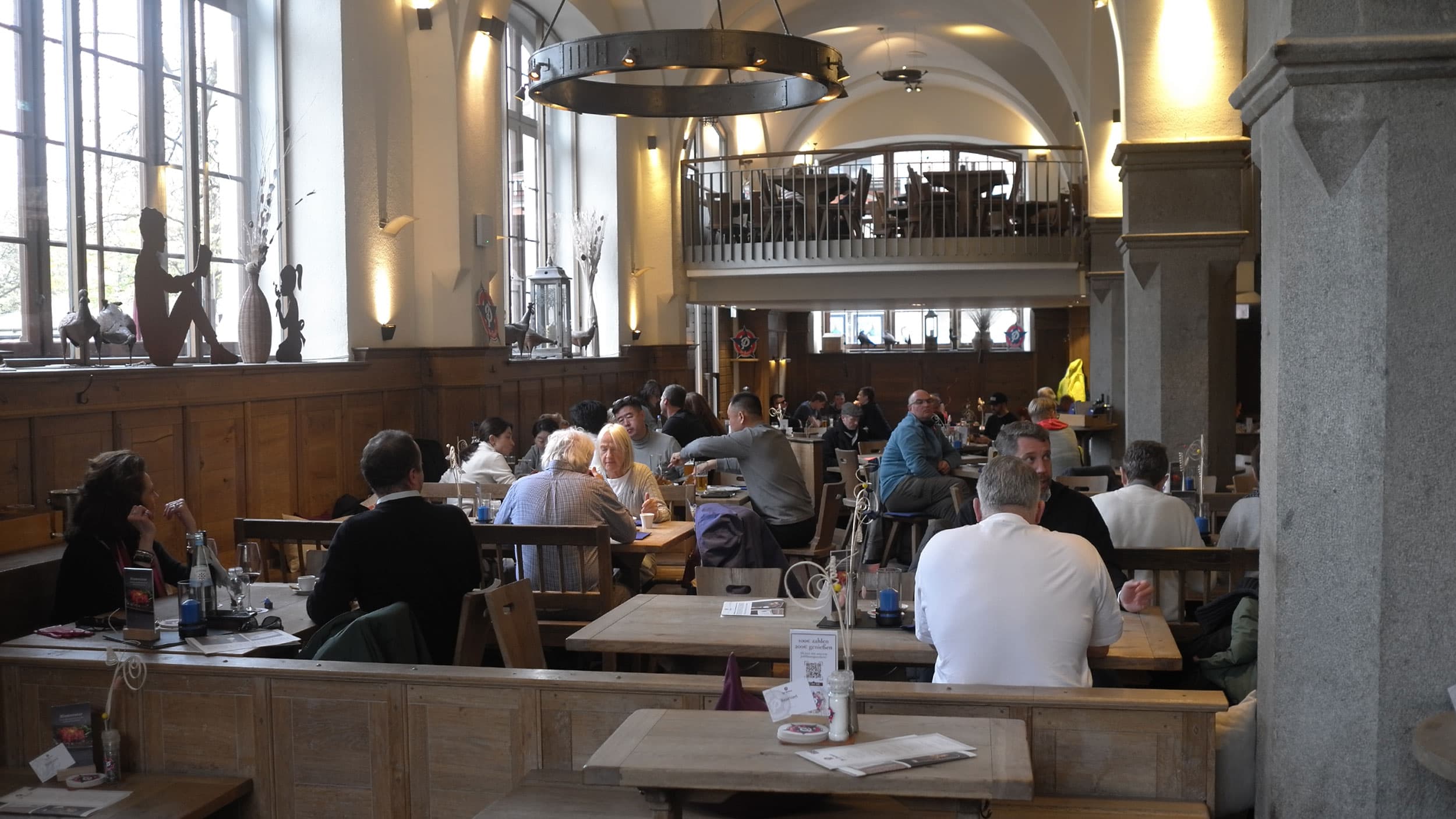 A spacious restaurant interior with light wood furnishings and large windows, filled with people seated at wooden tables. Decorative elements and a mezzanine level are visible in the background. The atmosphere is lively and casual.