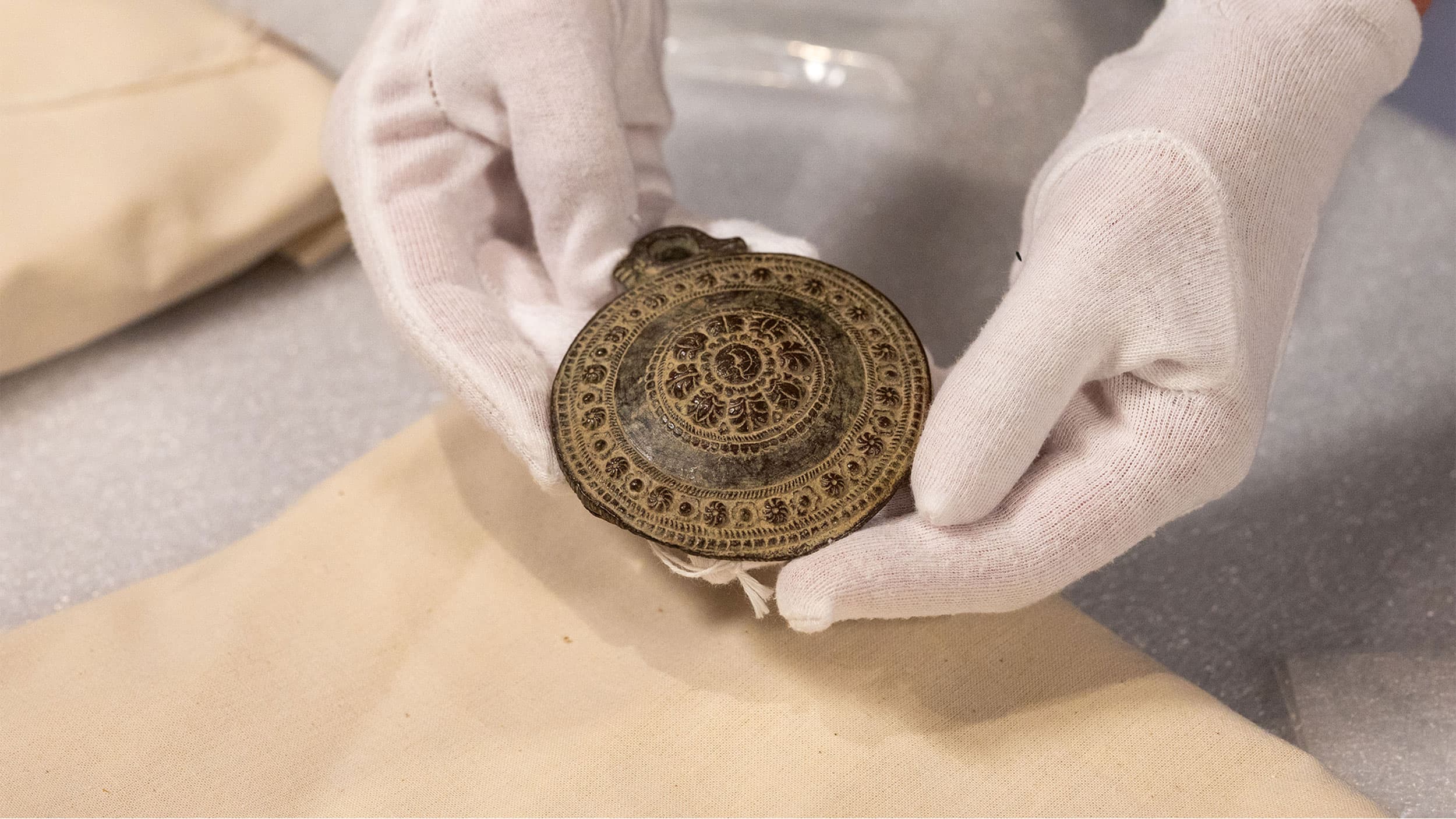 A person wearing white gloves holds an ornate circular artifact with intricate patterns carved into its surface.
